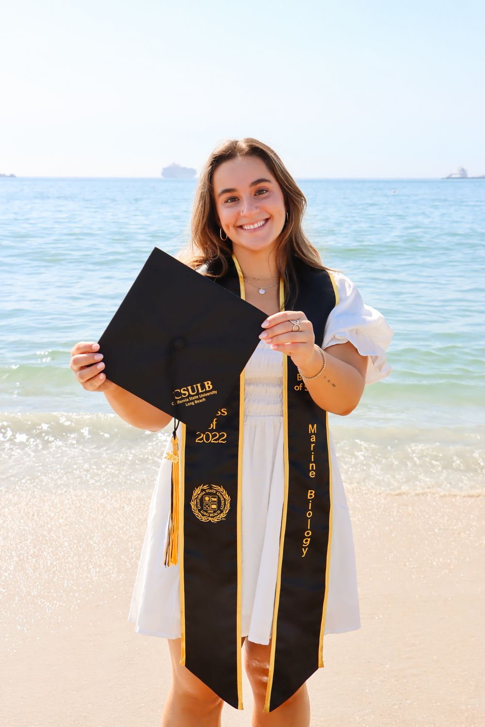 Graduate displays graduation stole and tassel against beach background.