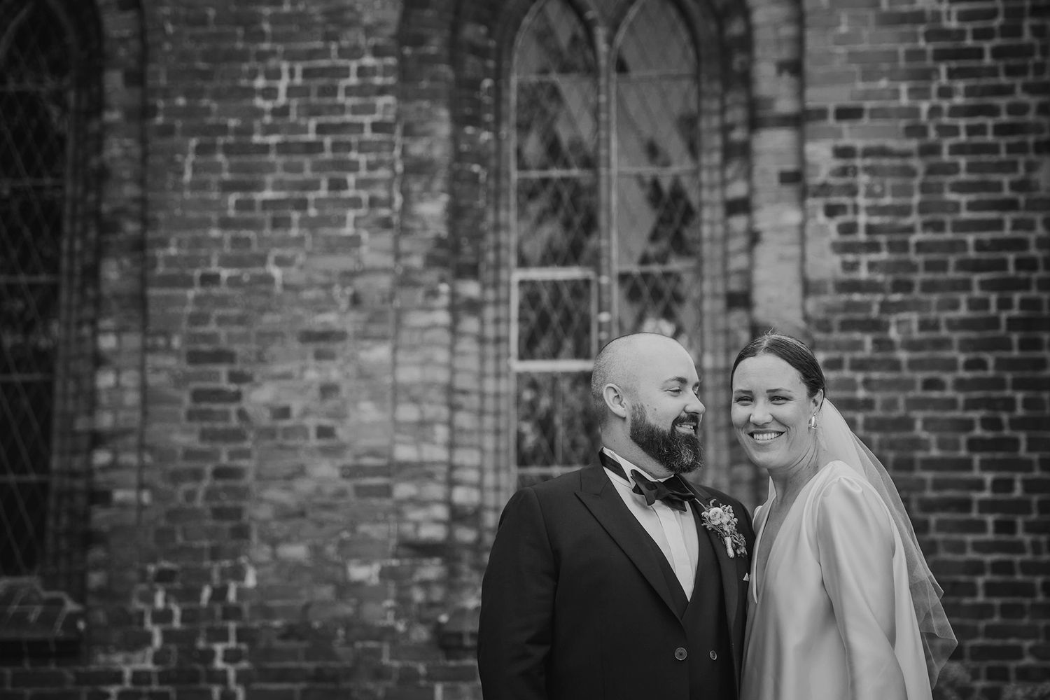 En brud og en brudgom poserer til et bryllupsfoto foran en murstenskirke med buede vinduer.