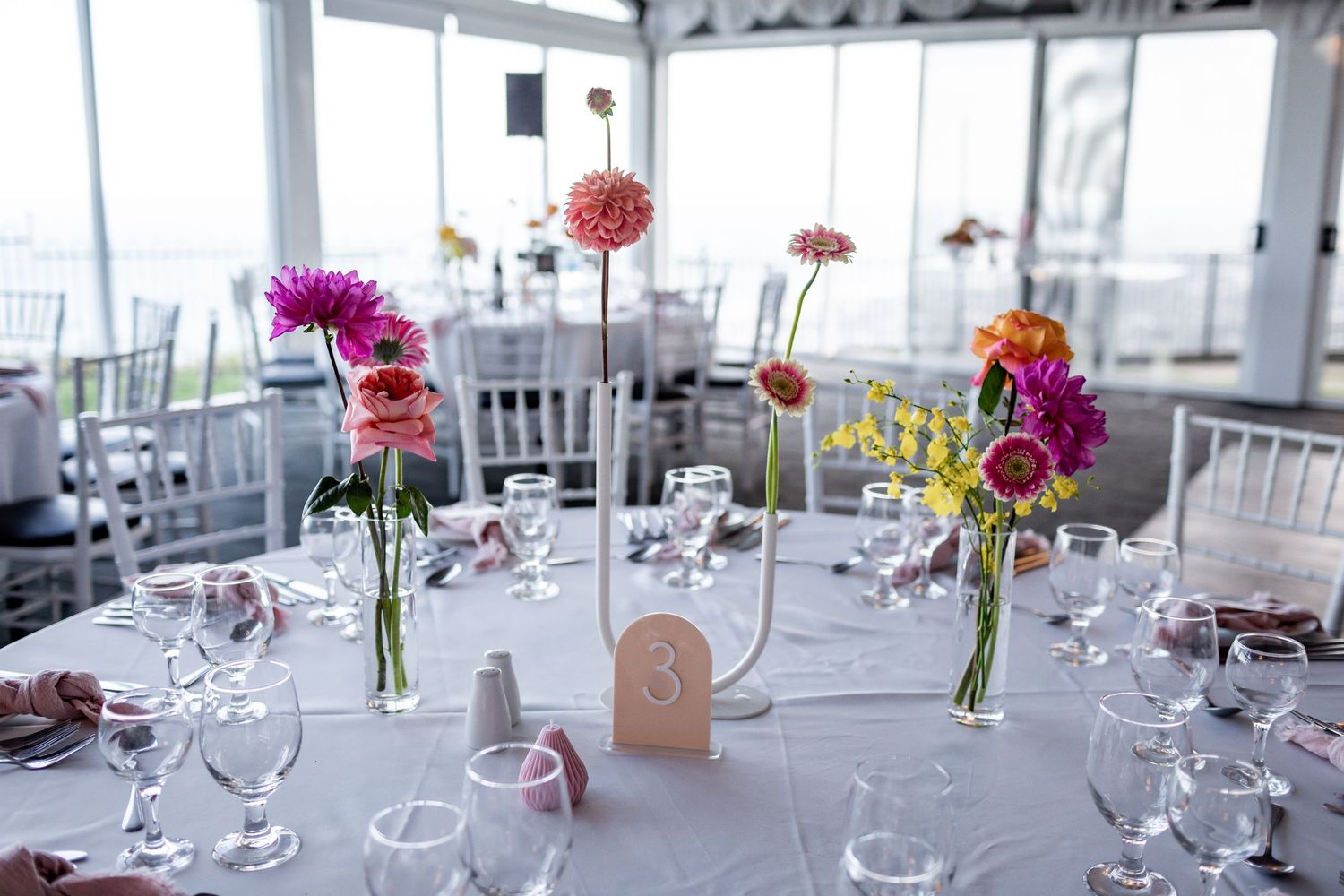 Multiple bud vases with colorful flowers arranged on a round table with wine glasses and place settings.
