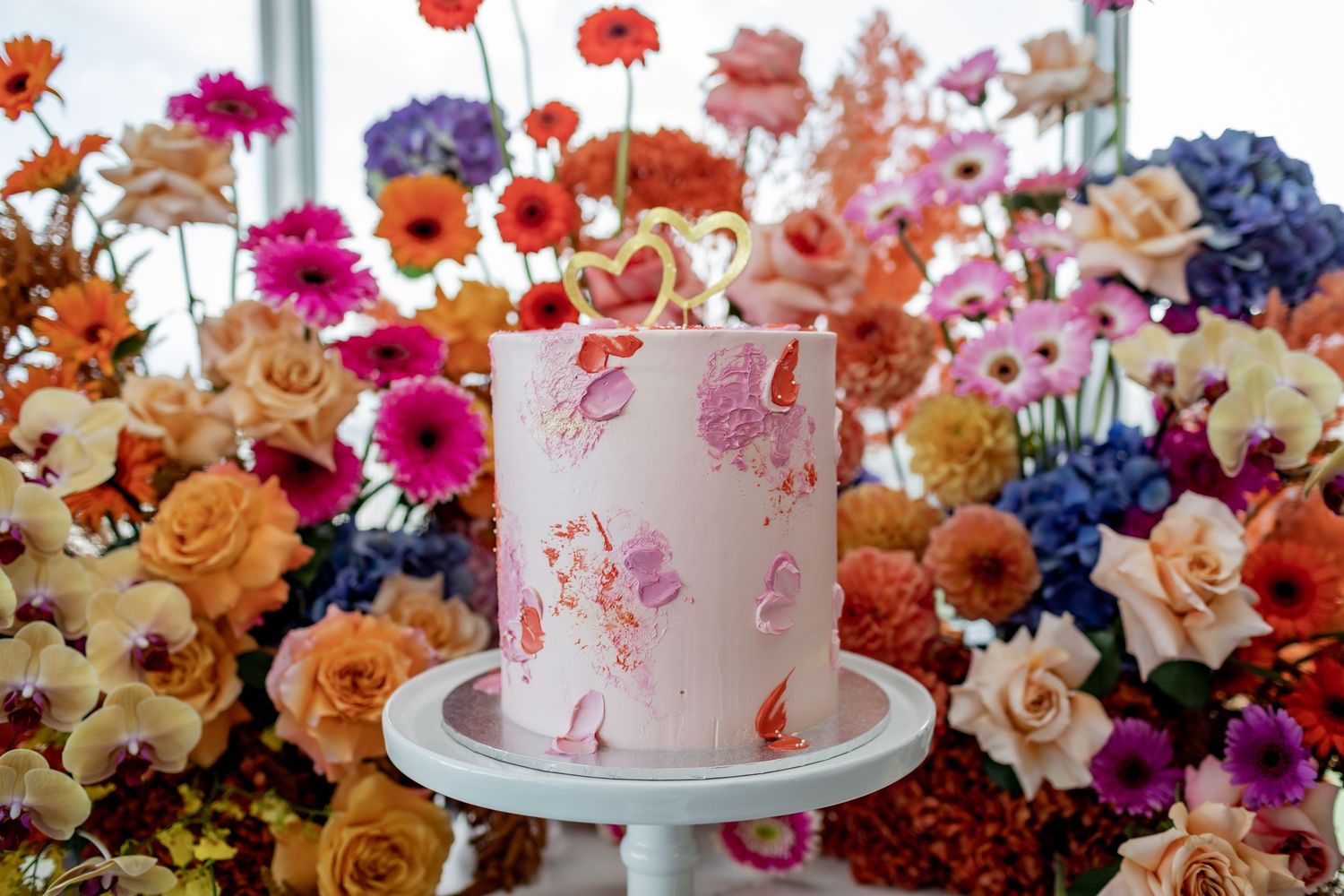 Wedding cake with pink watercolor design surrounded by vibrant mixed flowers on display stand.