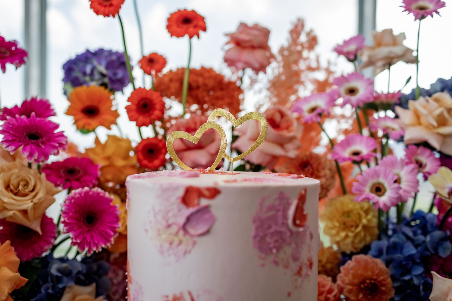 Close-up of pink wedding cake with floral design against colorful flower backdrop.