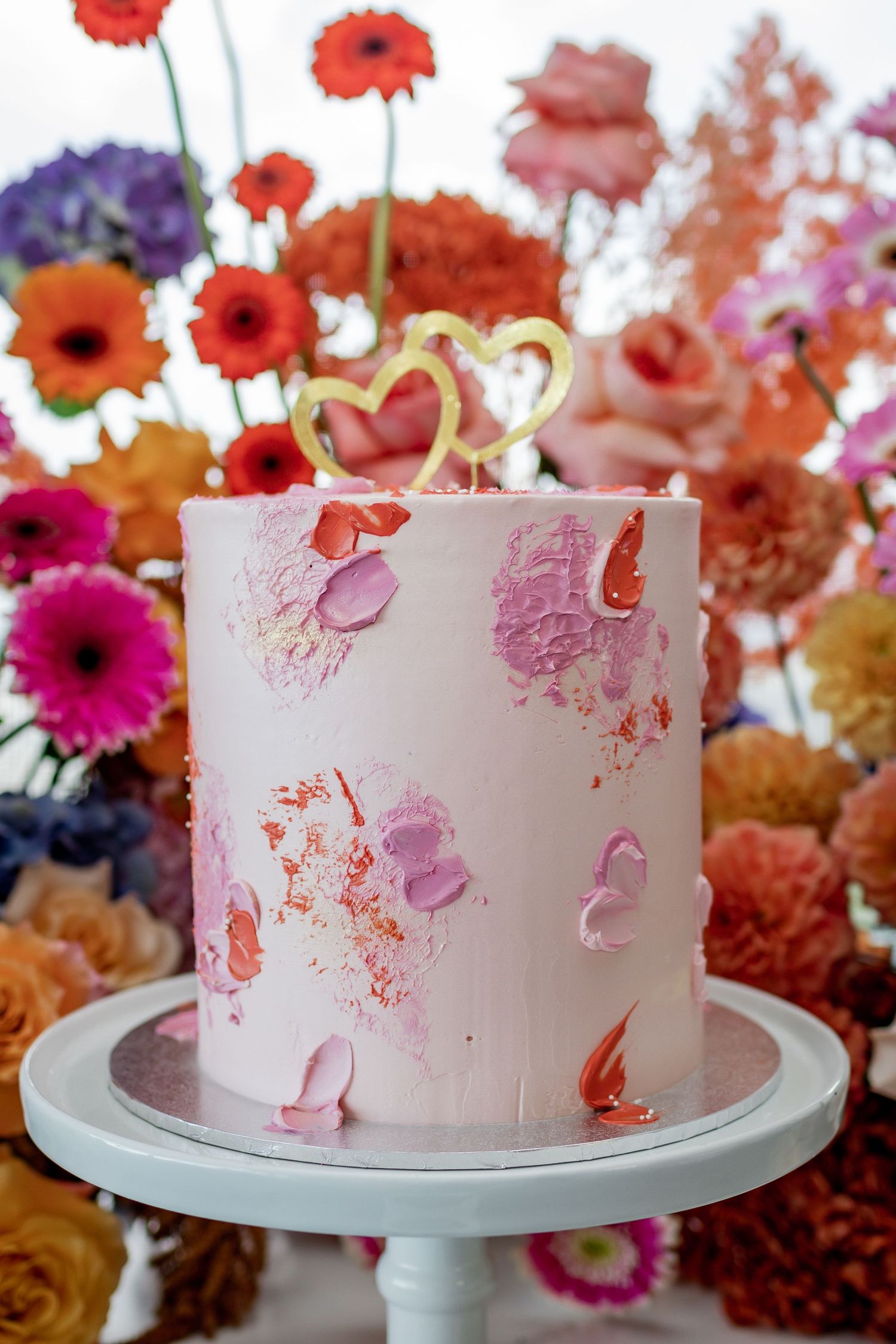 Pink wedding cake decorated with watercolor-style petals and gold heart topper on white stand.