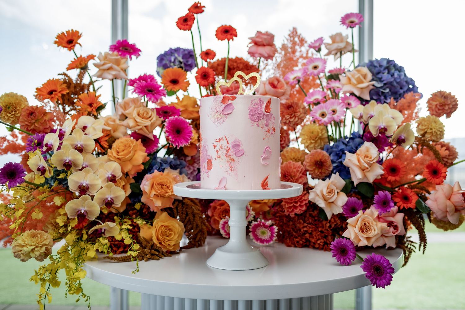 Pink wedding cake surrounded by lush colorful floral arrangements on white pedestal stand.