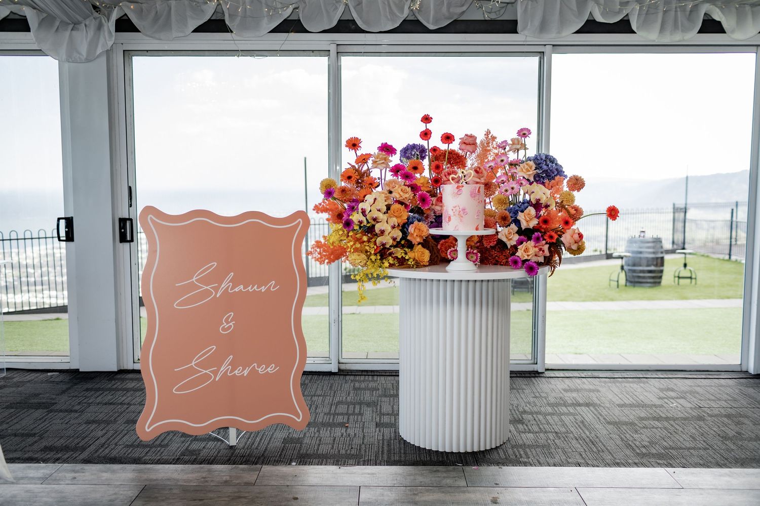 Large coral-colored welcome sign and floral arrangement in white column vase by window with ocean view.