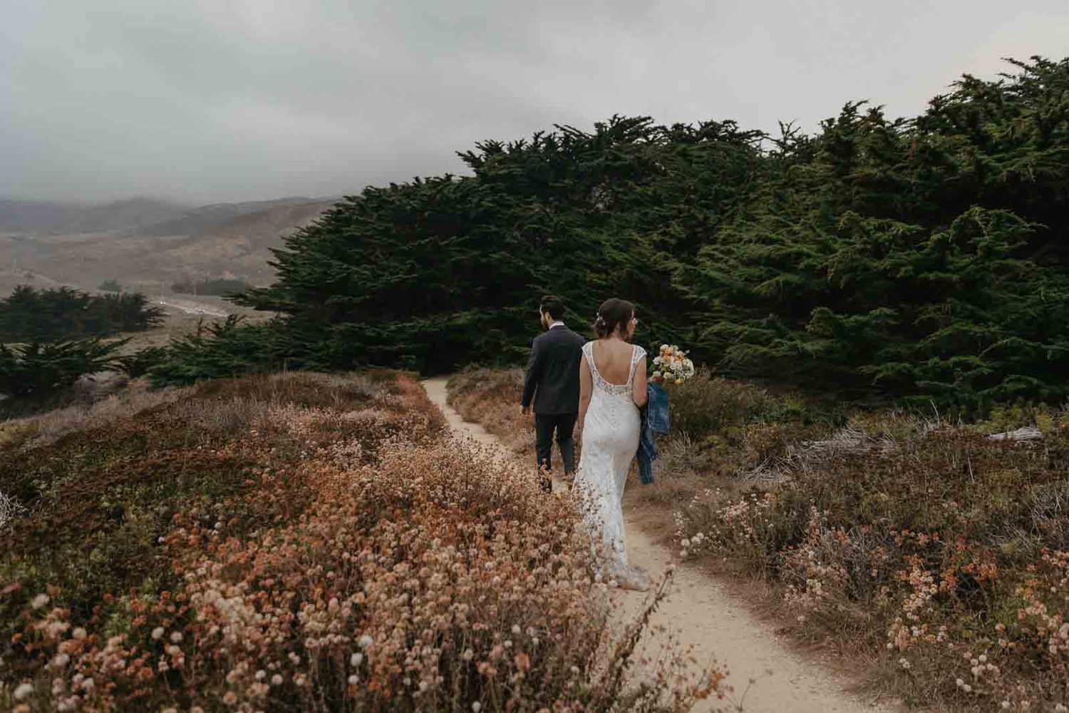 A newlywed couple walks along a coastal trail surrounded by cypress trees and fog.