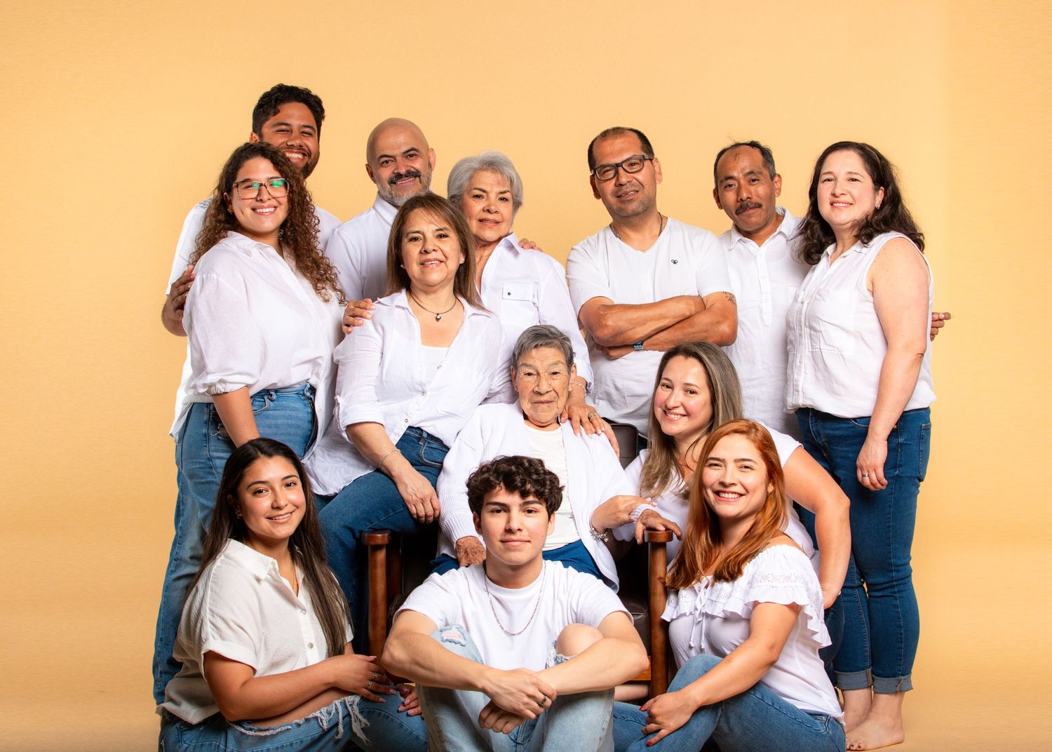 A large multigenerational Hispanic family poses together smiling in matching white tops on yellow background.
