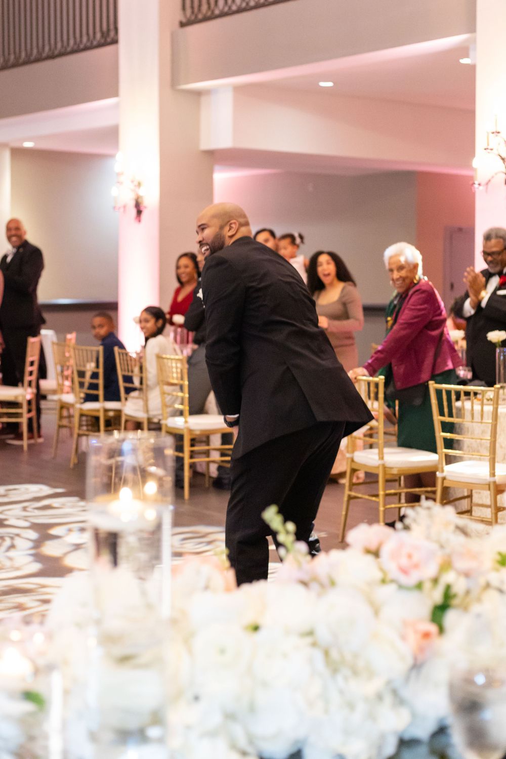 Reception guests watch as someone dances energetically at an elegant wedding venue with floral centerpieces.