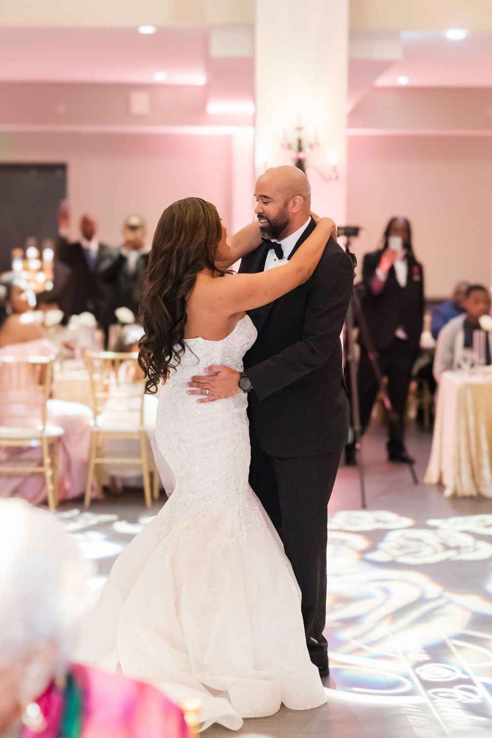A couple embraces during their wedding dance in a pink-lit ballroom with geometric floor patterns.