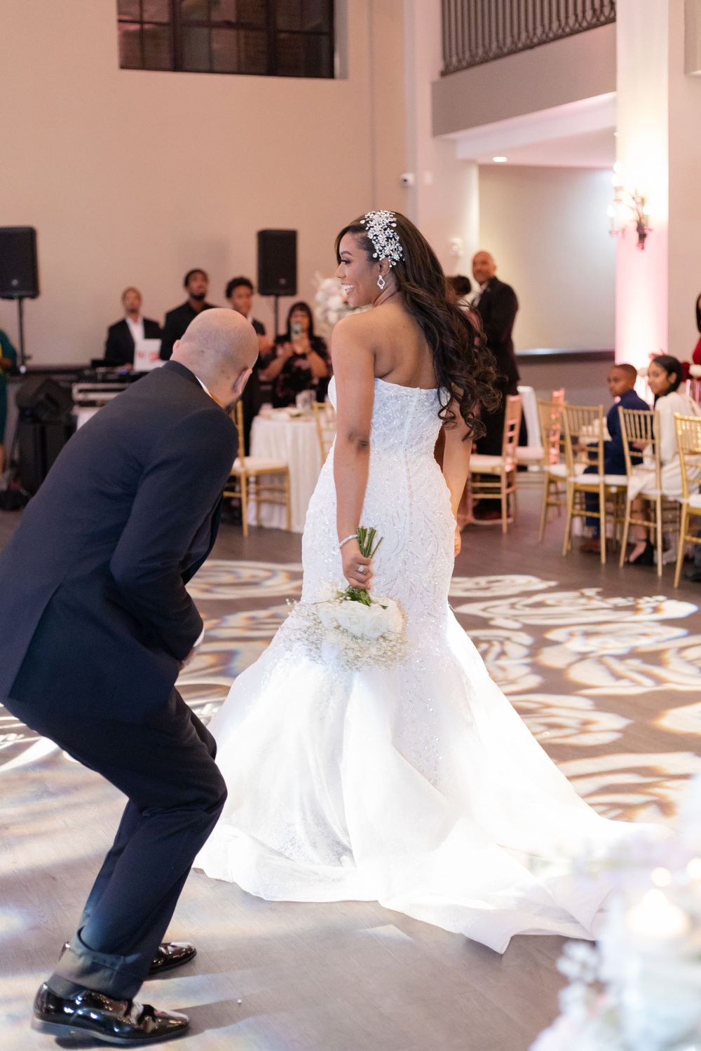 A bride in a mermaid wedding gown dances at her reception while guests look on from gold chairs.