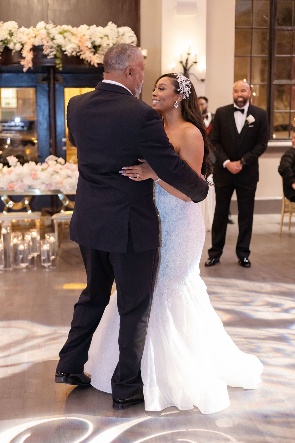 Two people share an intimate wedding dance while a guest in a black tuxedo watches in the elegant ballroom.