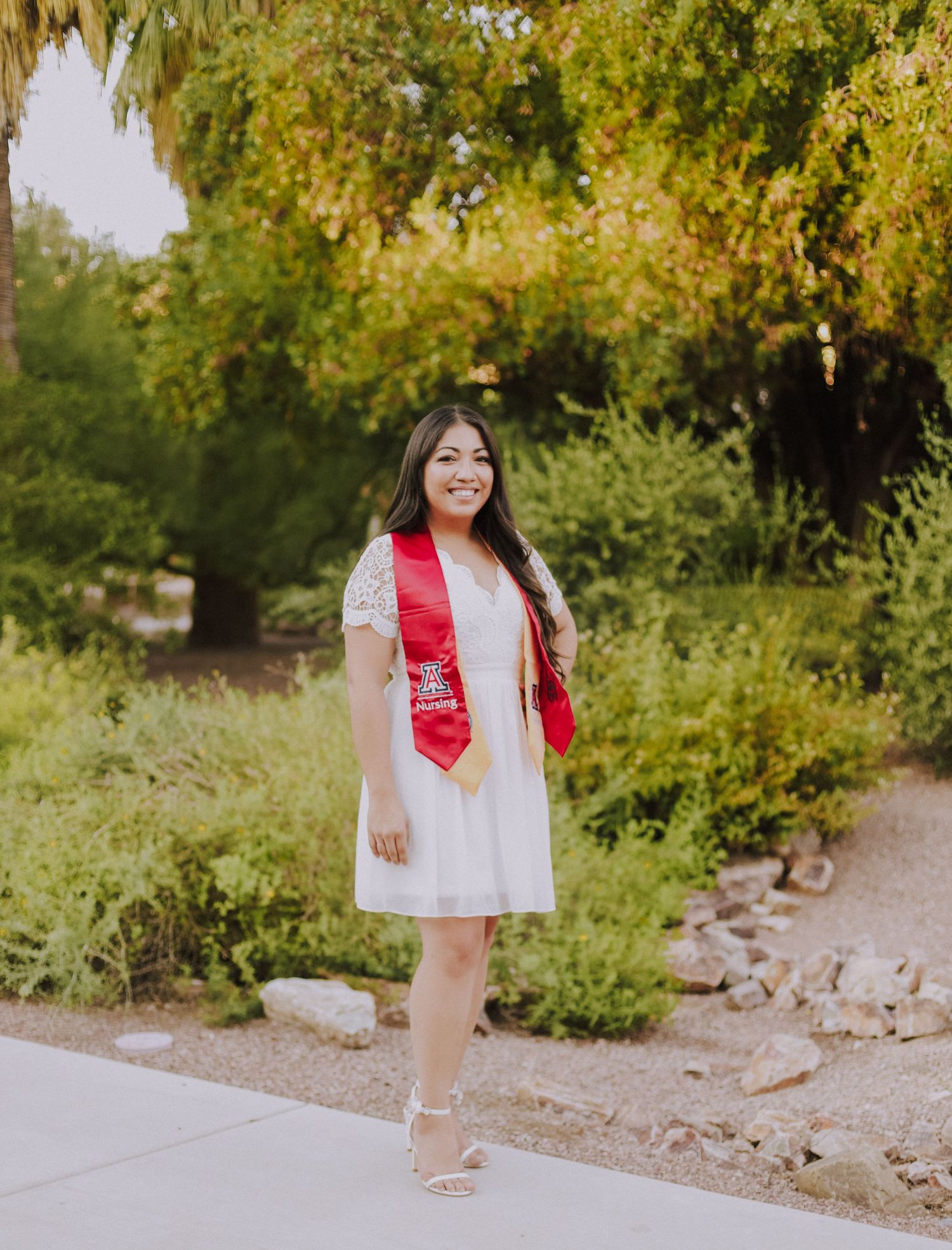 A graduate in a white dress and red stole poses outdoors with palm trees and greenery in background.