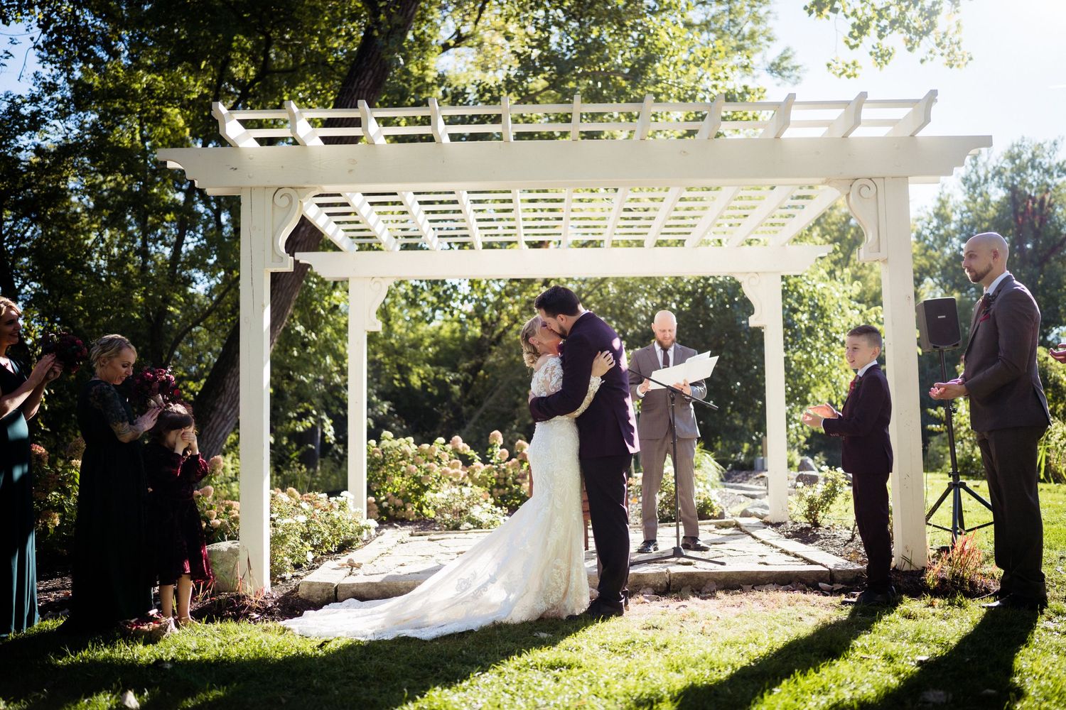 Wedding ceremony under white pergola in sunlit garden with guests watching.