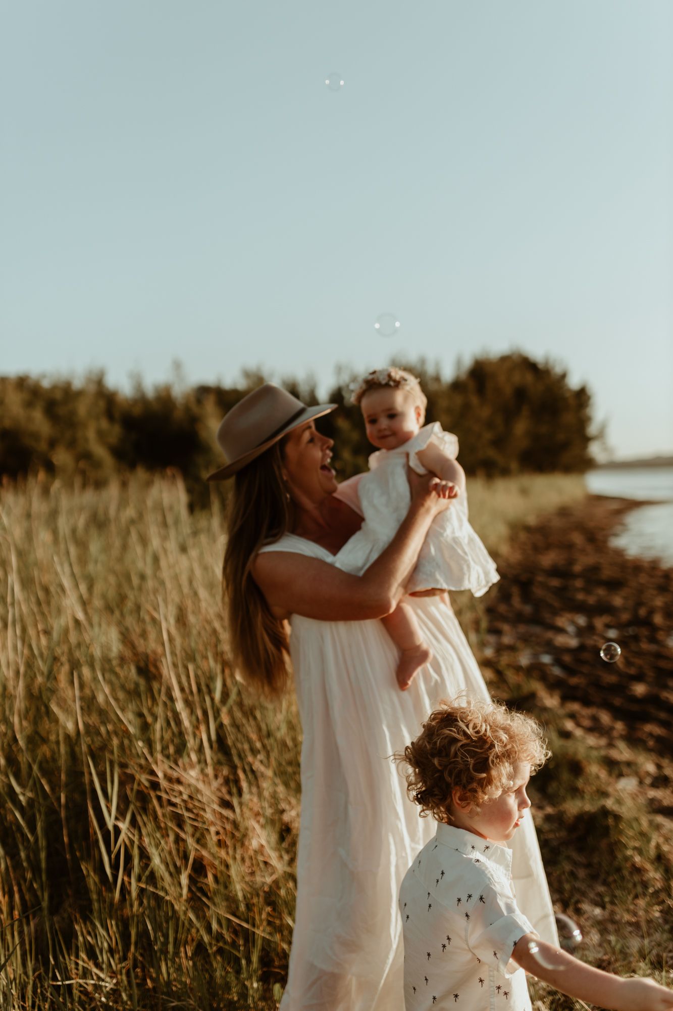 A woman in a hat and white dress holds two small children in a seaside field at sunset.