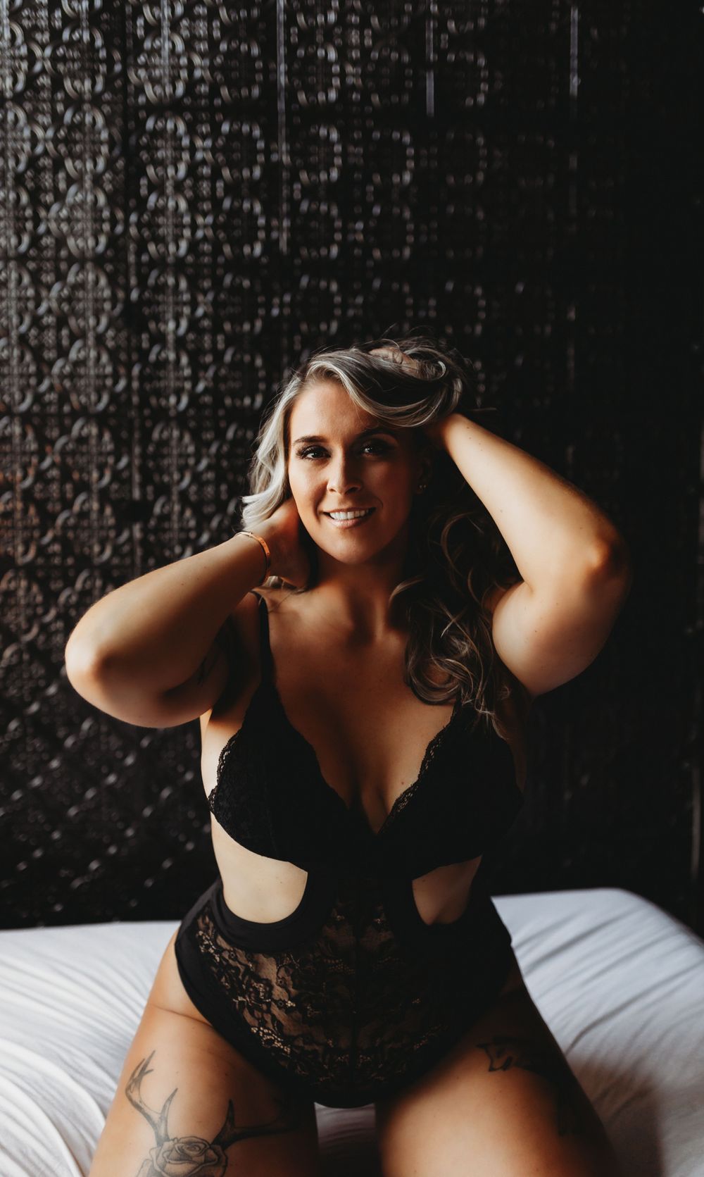 A woman in black lace lingerie poses confidently on white bedding against a dark patterned wall.