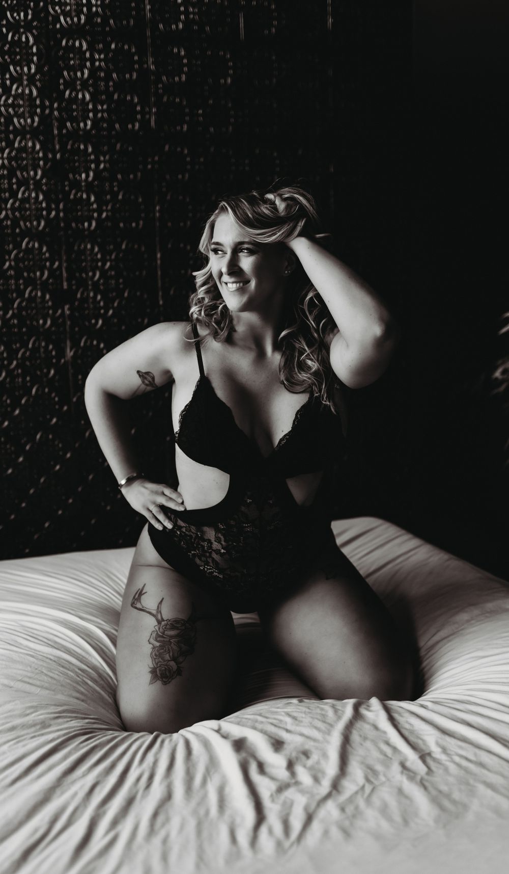 Black and white boudoir image showing a person in lingerie posing confidently on a bed in dramatic lighting with dark backdrop.