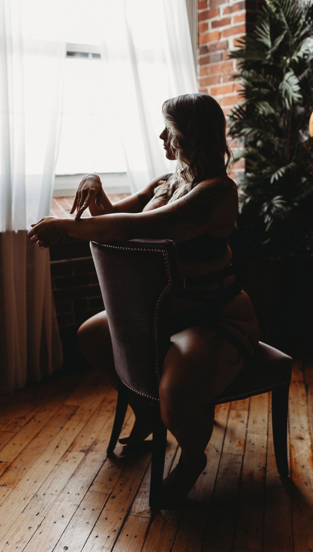 A silhouetted figure sits pensively in an upholstered chair by a window with sheer curtains and houseplants.