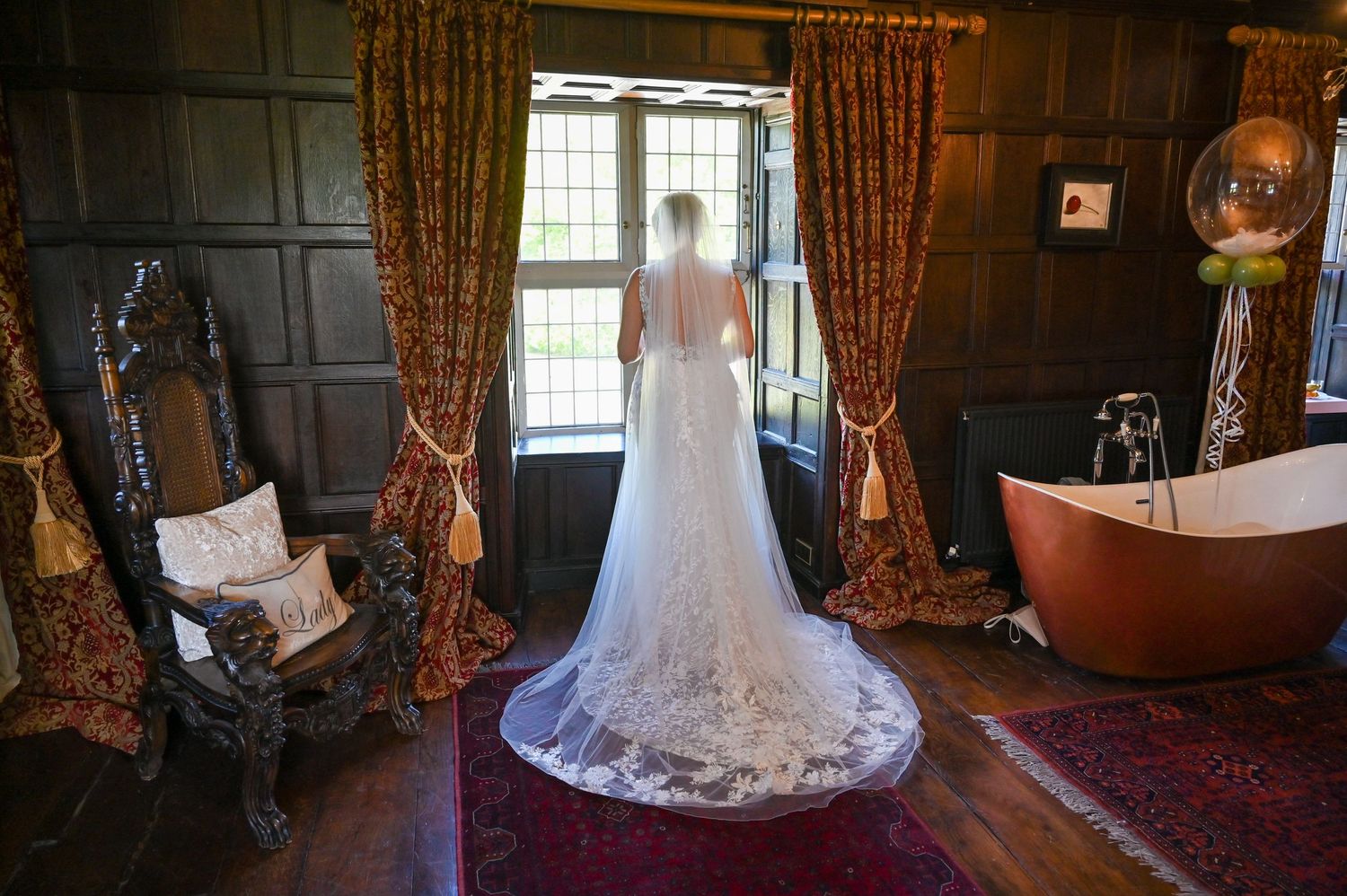 A wedding dress with a flowing lace train photographed from behind in an elegant wood-paneled room with vintage decor.