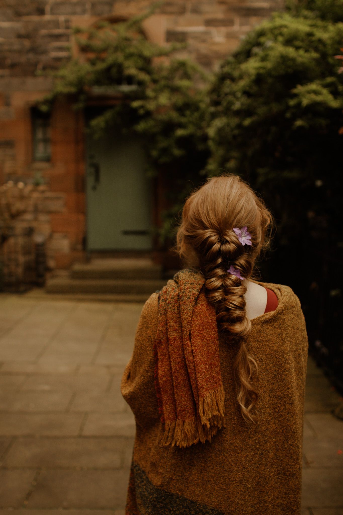 Back view of braided hair with purple flowers against rustic stone wall.