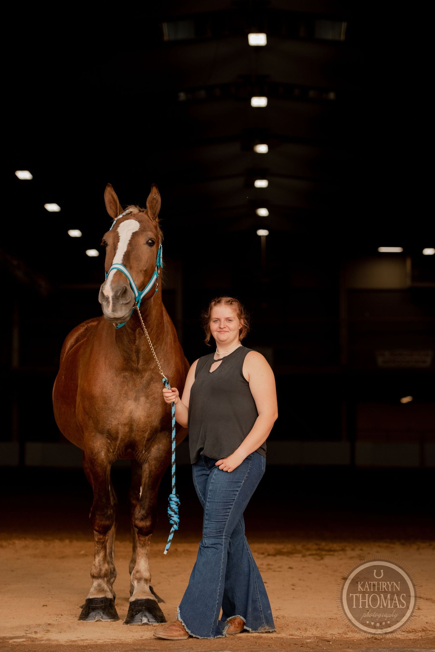 A bay horse wearing turquoise tack stands with its handler in a dimly lit indoor arena.