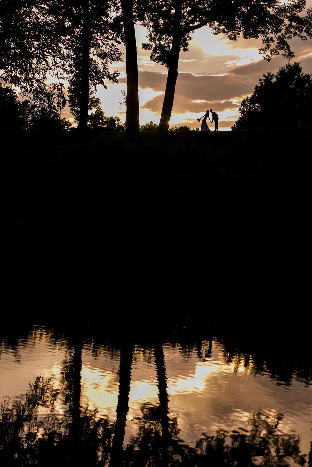 A serene lakeside sunset with silhouetted trees reflecting on calm waters.