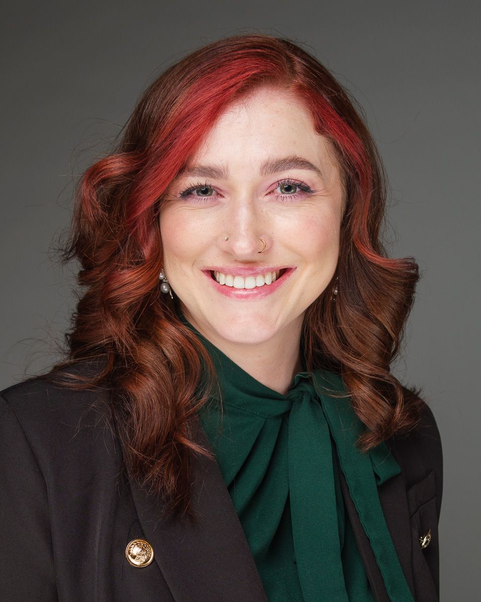 Professional headshot of a smiling person with burgundy and brown hair wearing a black blazer and emerald green blouse.