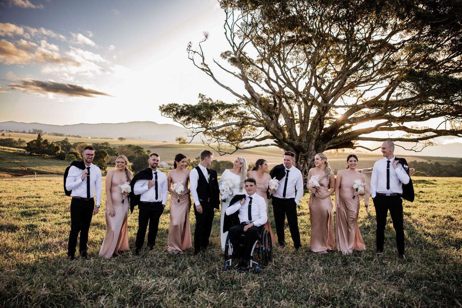 Wedding party poses at sunset under a large tree on a hilltop with dramatic sky.