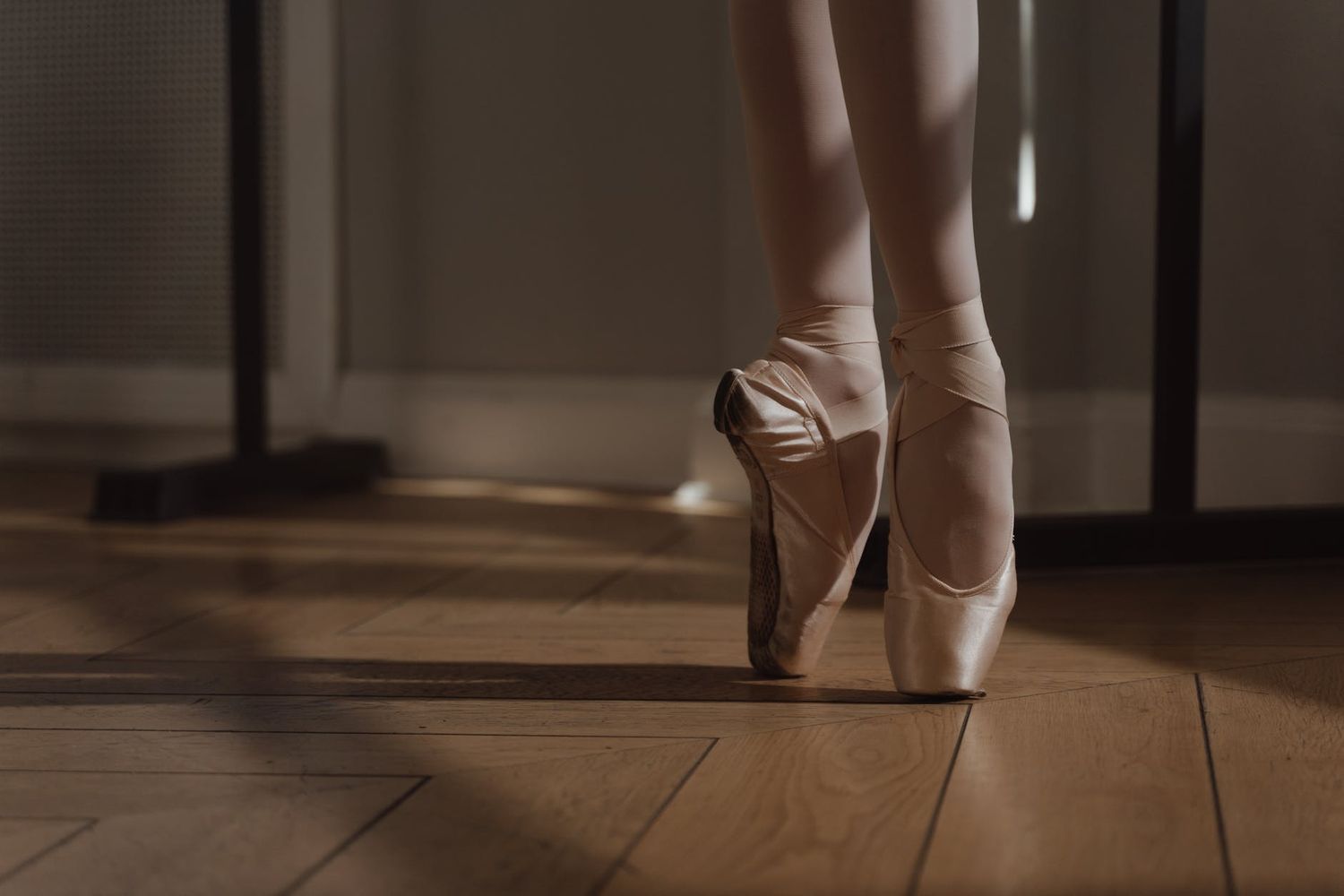 Close-up of pink satin pointe shoes on hardwood floor in dim lighting.