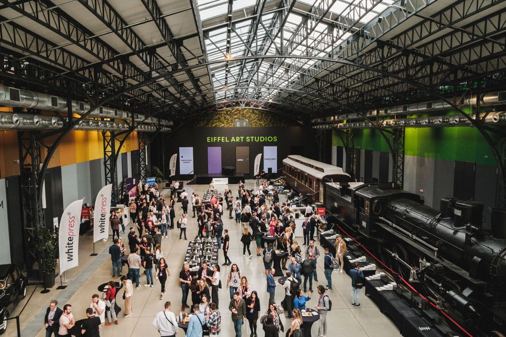 Crowded conference hall with high glass ceiling and industrial architecture filled with attendees.