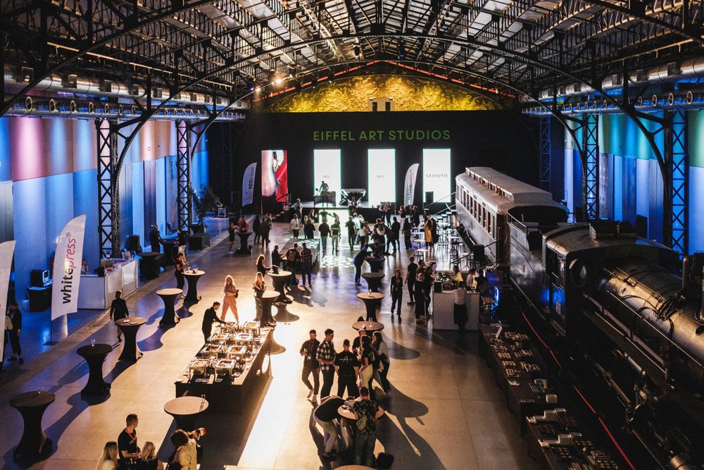 Large industrial venue space with crowds mingling under colorful lighting and high ceilings.