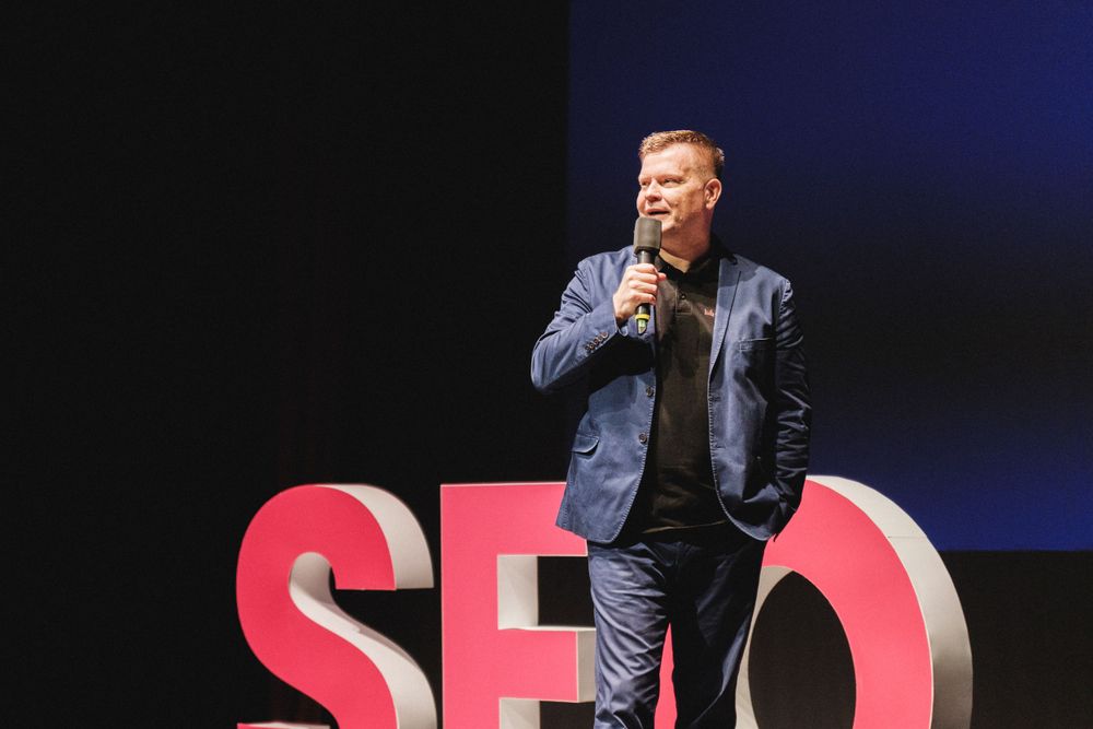 A speaker presents on stage with large red SEO letters in the background.