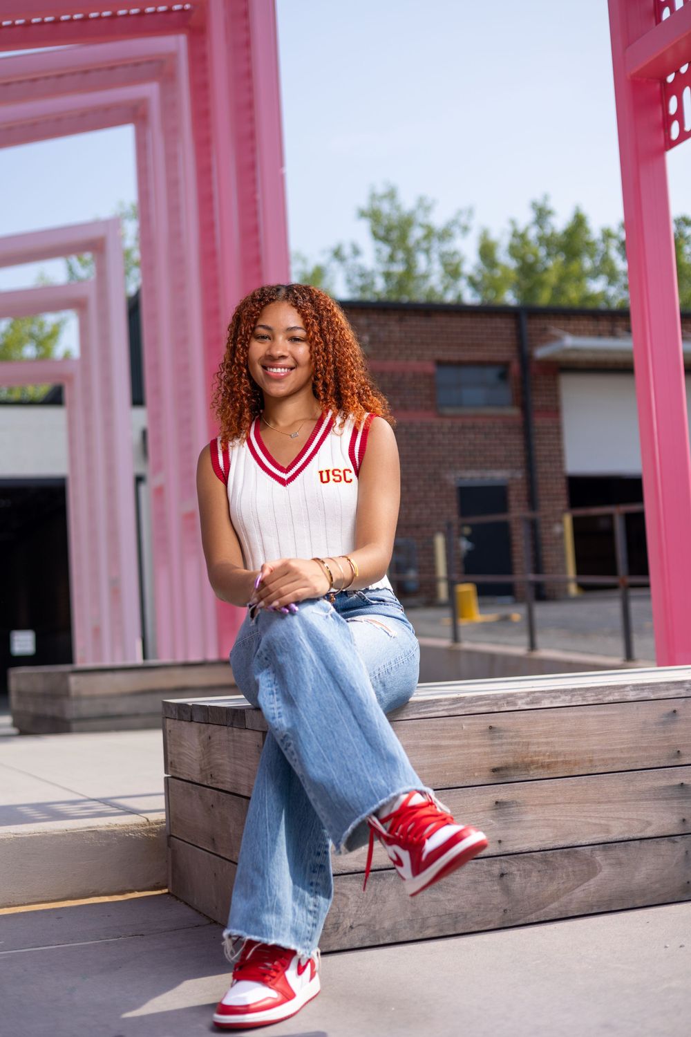 Relaxed pose on wooden bench wearing vintage-style outfit with red sneakers and pink archway backdrop.