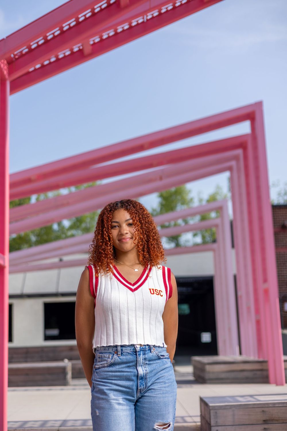 Casual outfit featuring white varsity tank top and jeans posed under pink architectural frames.