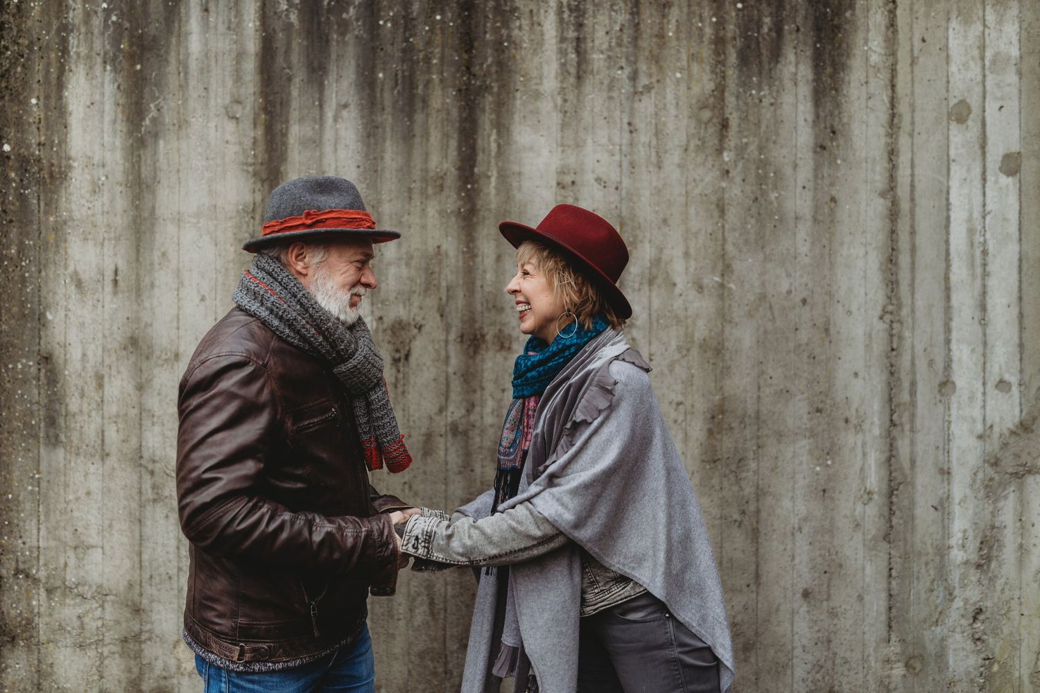 Two people in winter coats and hats share a laugh against a concrete wall.
