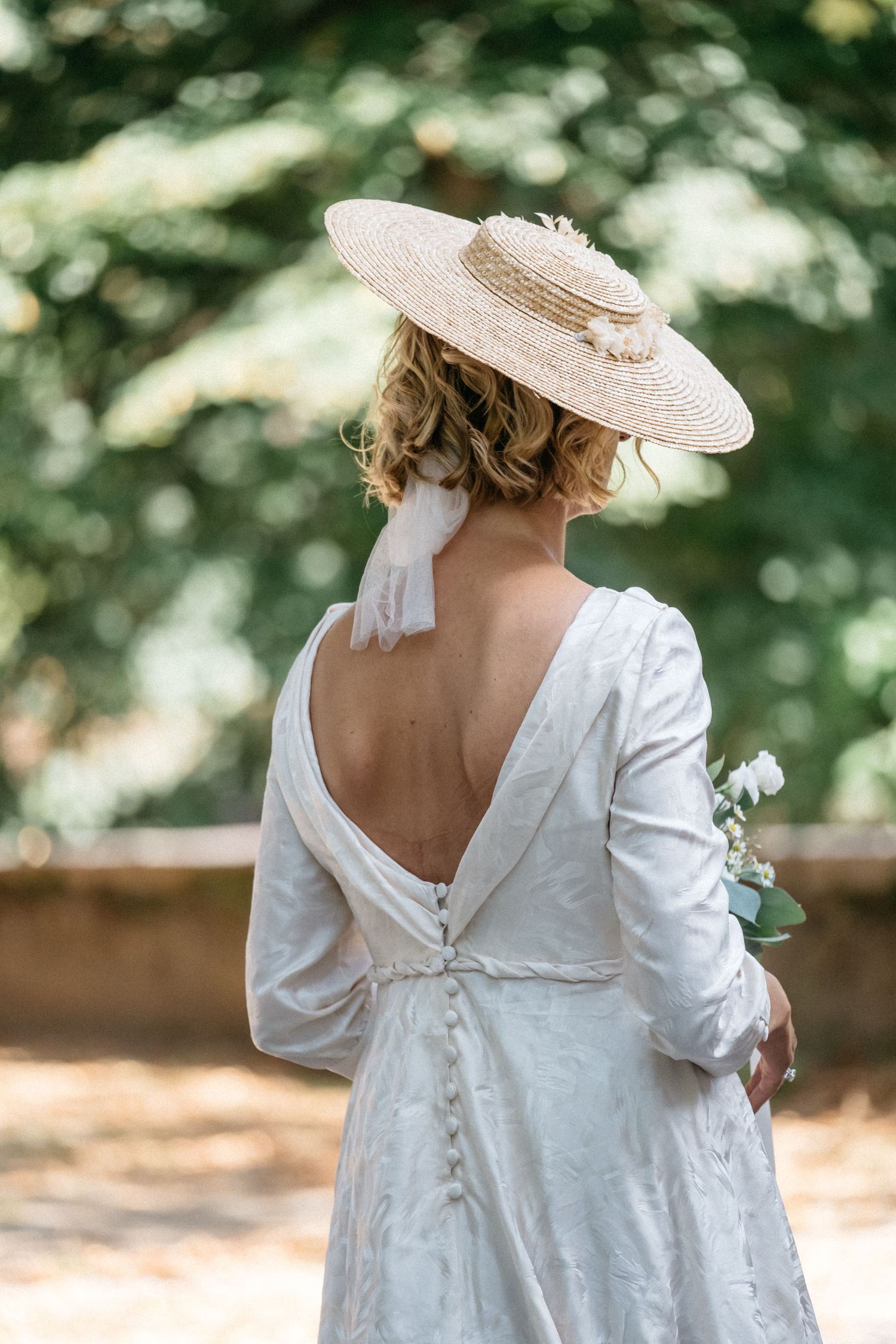 Back view of woman in vintage white dress with low back and straw hat standing in garden with bokeh background.