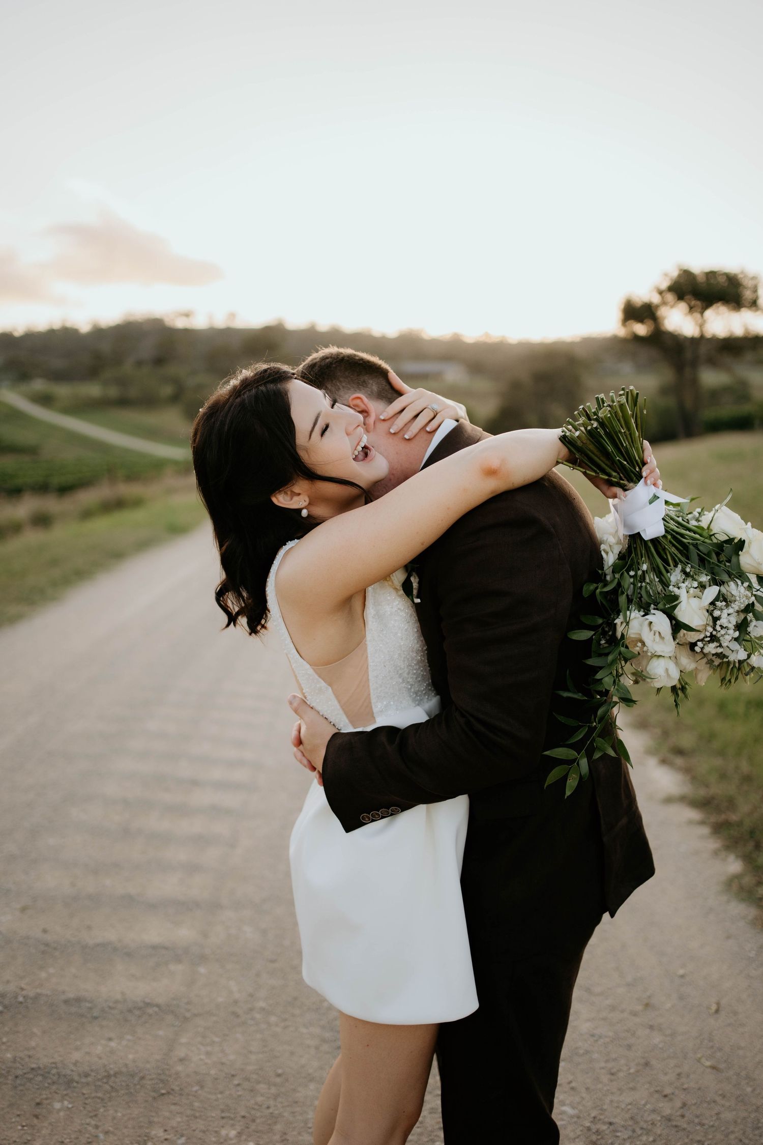 A couple shares a romantic embrace on a dirt road at sunset holding a white floral bouquet.