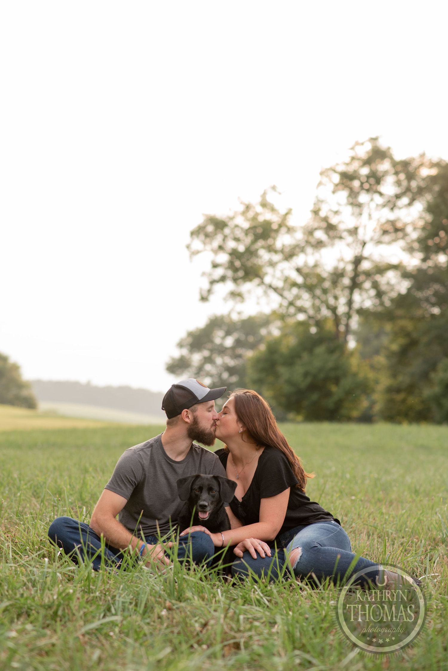 Couple sits together on grass with their black dog during sunset engagement photo session.