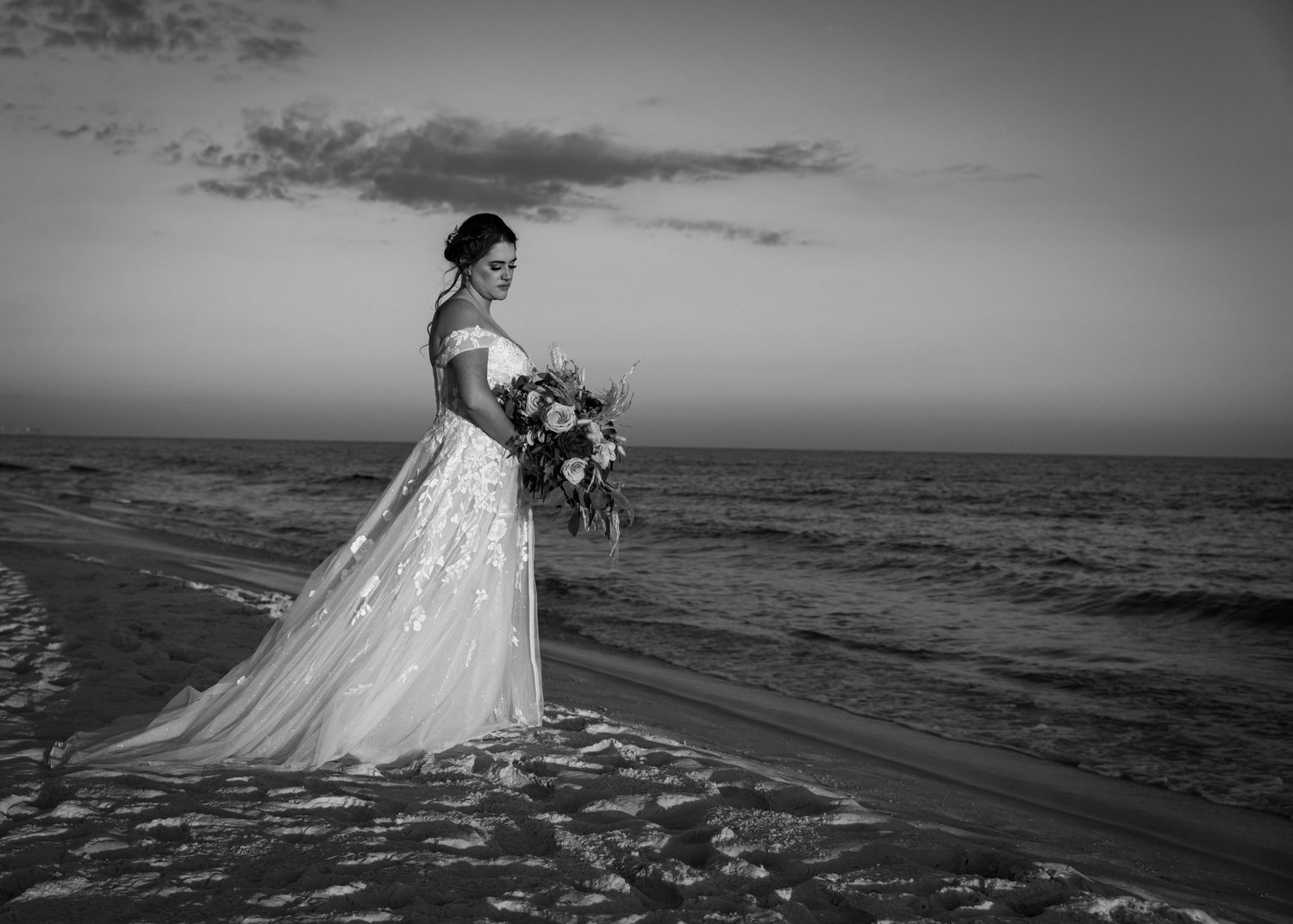 A black and white portrait of a bride holding bouquet on beach with ocean backdrop.