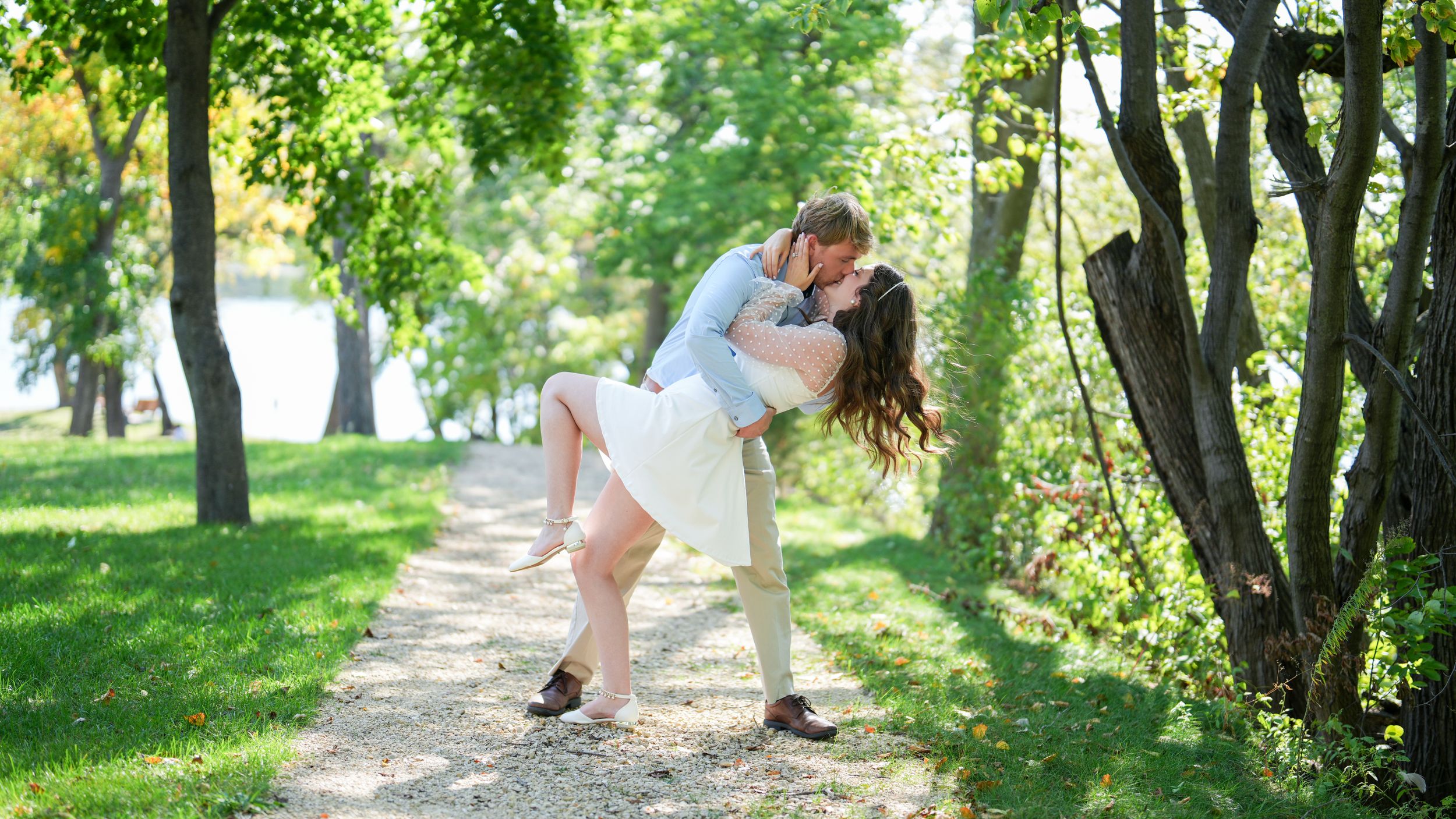 Couple sharing a romantic dip kiss on a sunlit garden path lined with green trees.