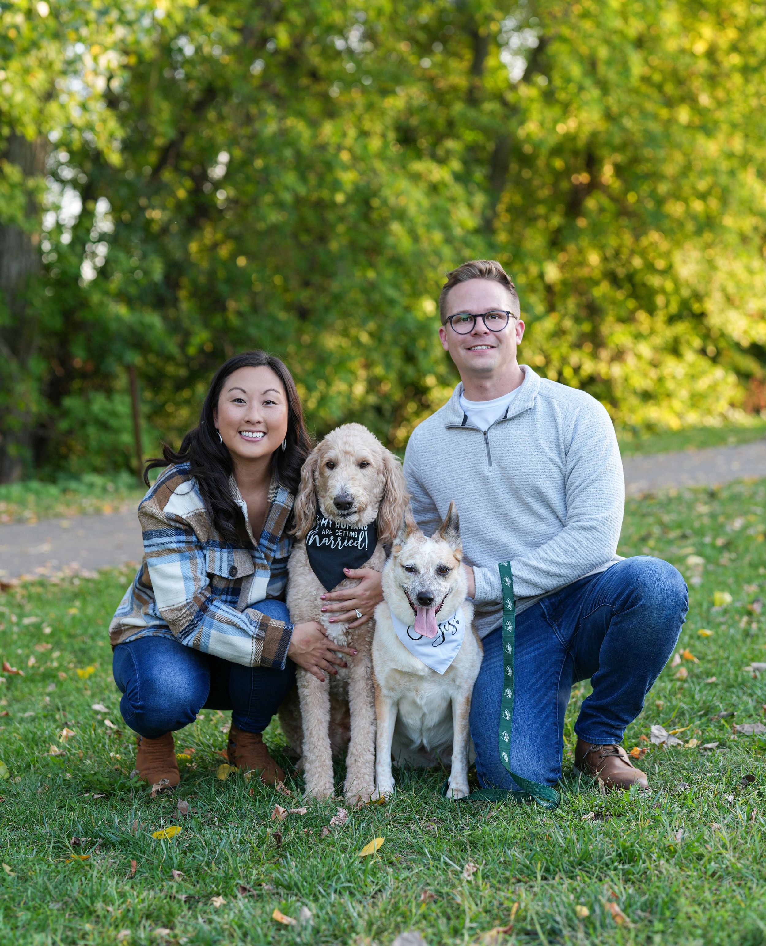 Smiling couple crouching with two dogs in a lush green park during fall engagement photos.