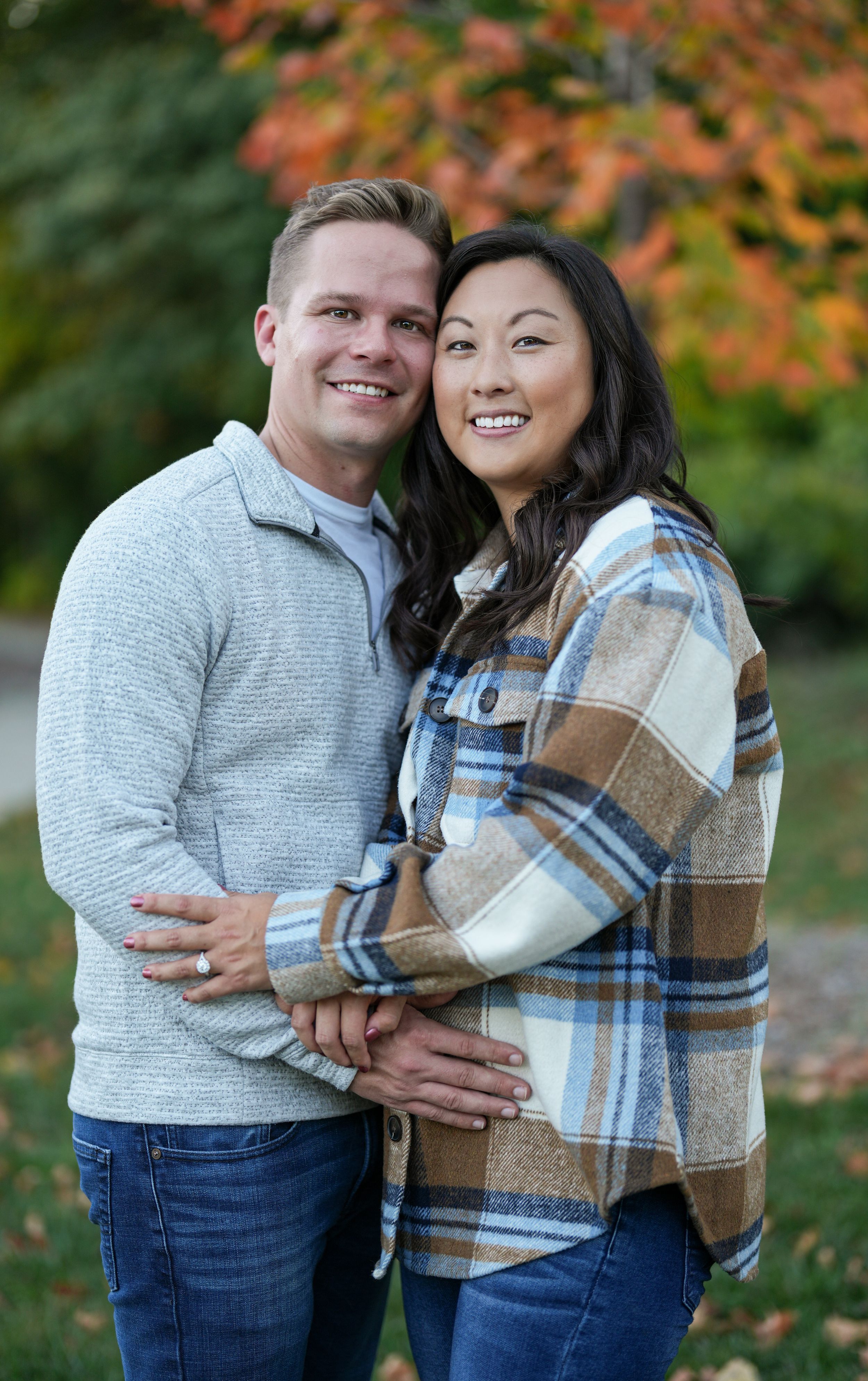 Smiling couple embracing outdoors with vibrant fall foliage in a warm autumn engagement photo.