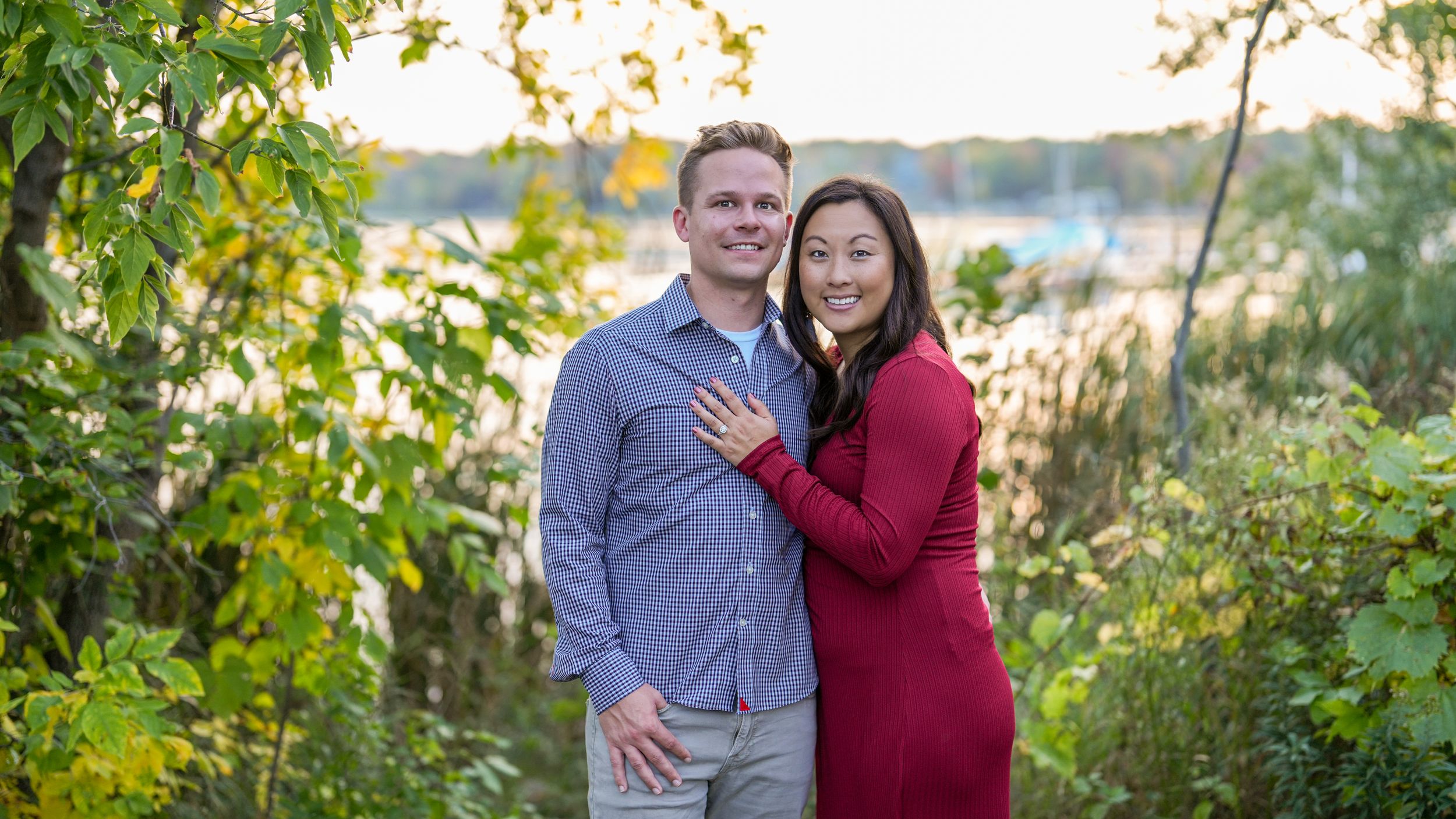 Engaged couple embracing near a lakeside with colorful autumn foliage in the background.