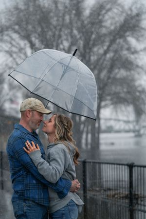 Couple embracing and smiling under clear dome umbrella in snowy weather by waterfront.