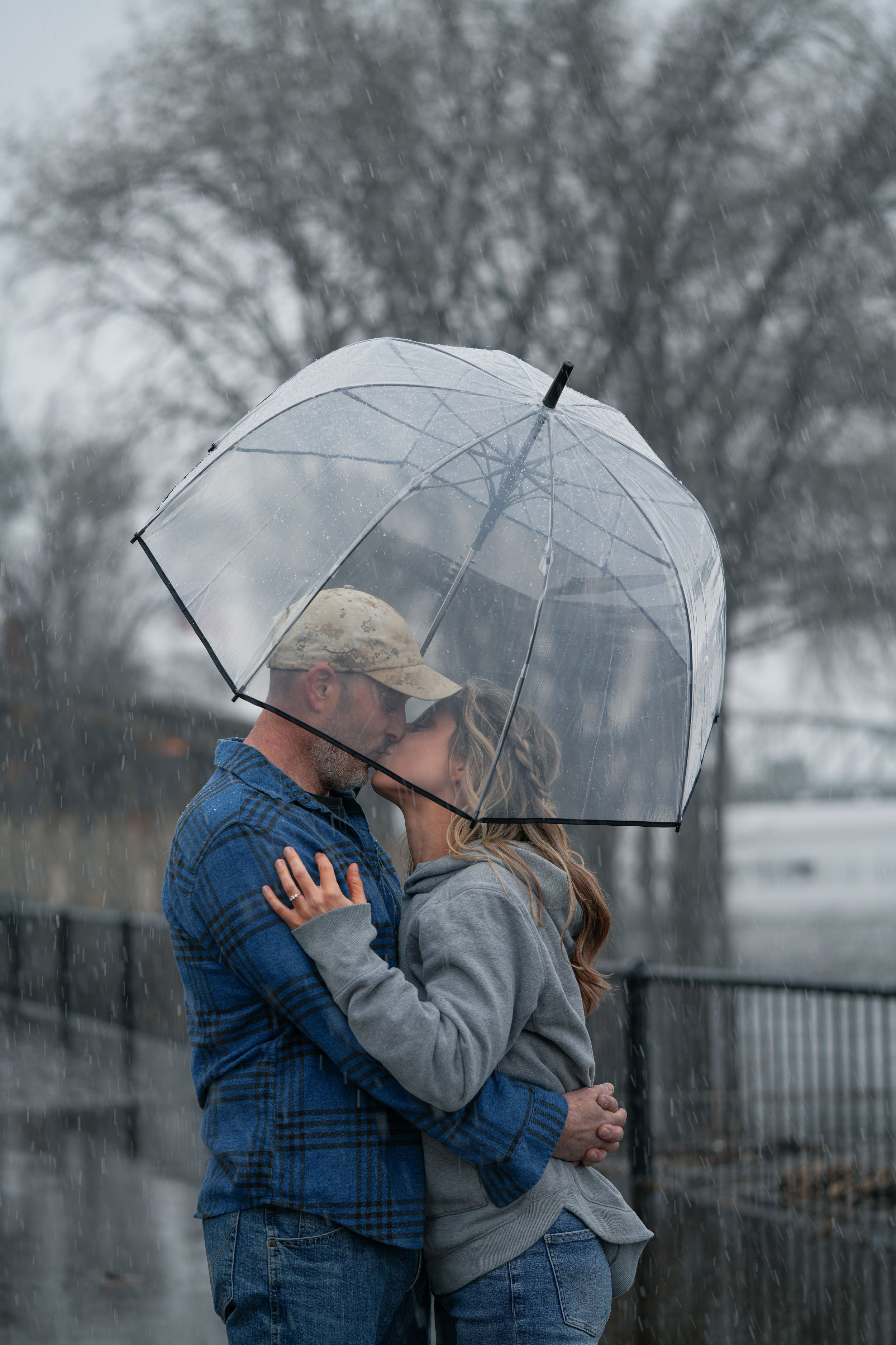 Romantic couple kissing under clear bubble umbrella in snowy winter rain by waterfront.