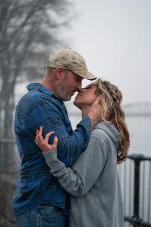 Couple sharing tender kiss in snowfall near river, man in plaid shirt and cap.