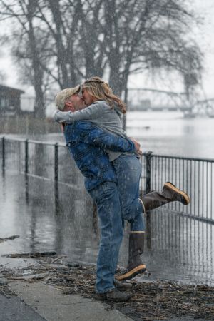 Man lifting woman in joyful embrace while kissing in heavy rain by flooded riverside.