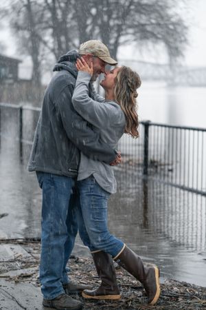 Couple kissing passionately in rain near flooded waterfront, woman wearing tall brown boots.