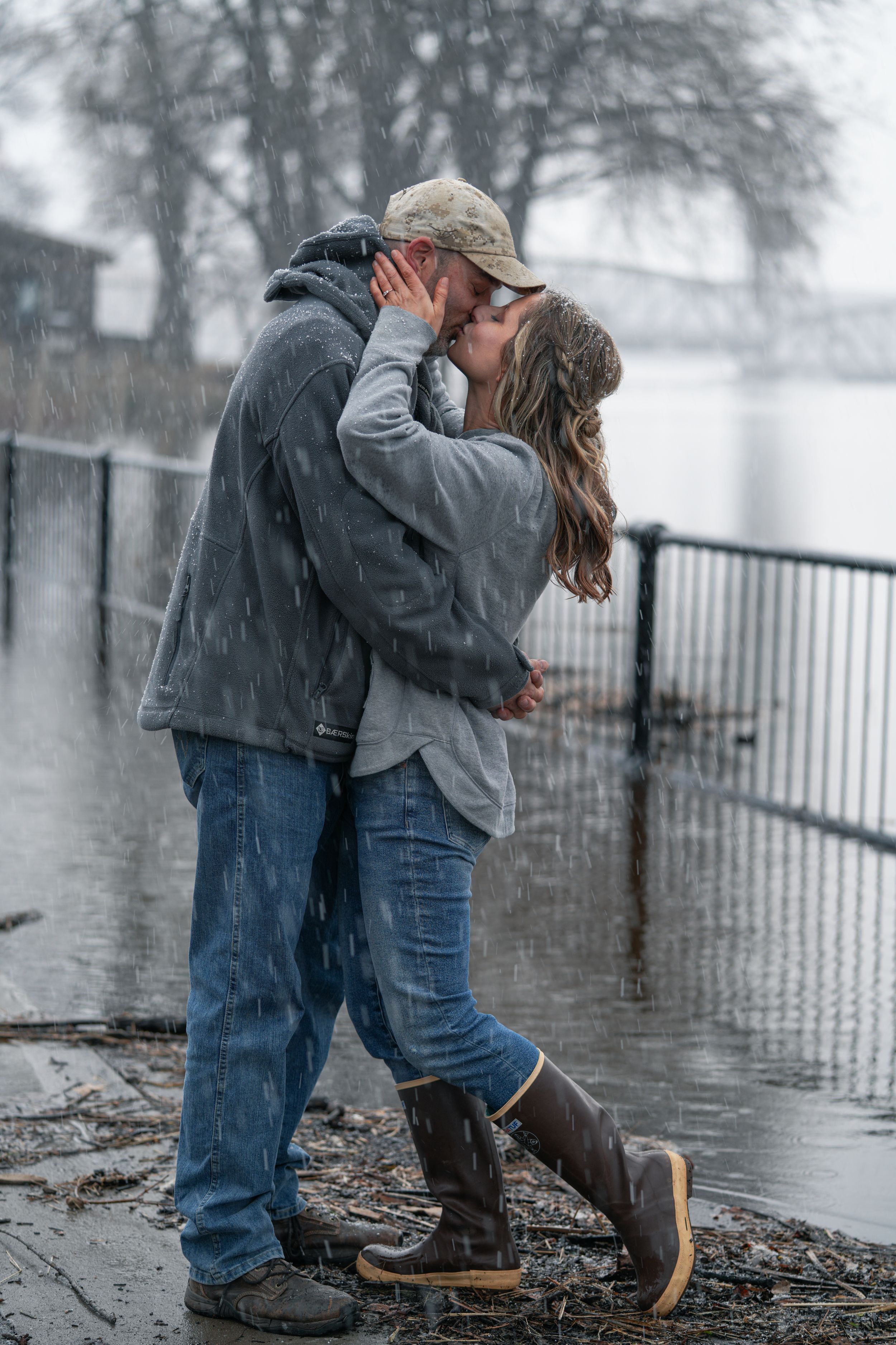 Couple sharing passionate kiss in heavy snowfall by riverside fence wearing casual winter clothes.