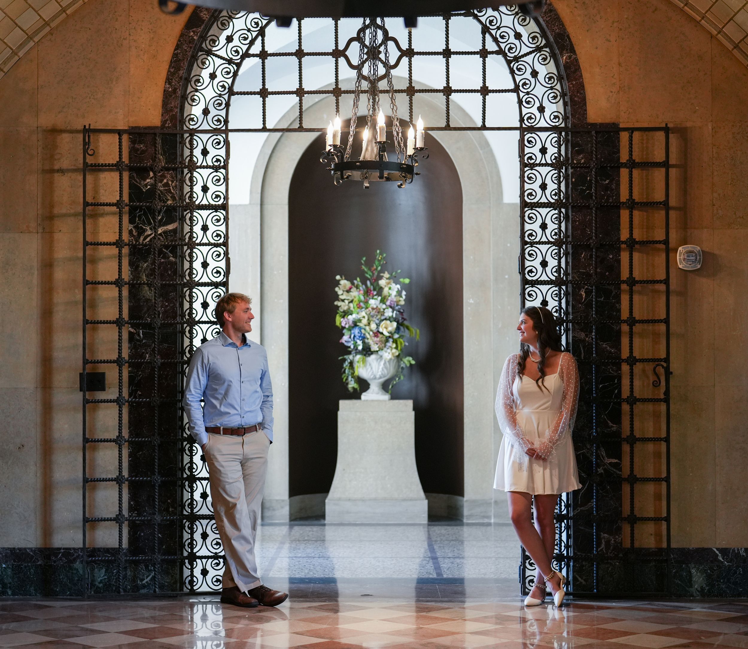 Elegant couple standing apart in ornate arched doorway with chandelier and floral arrangement.