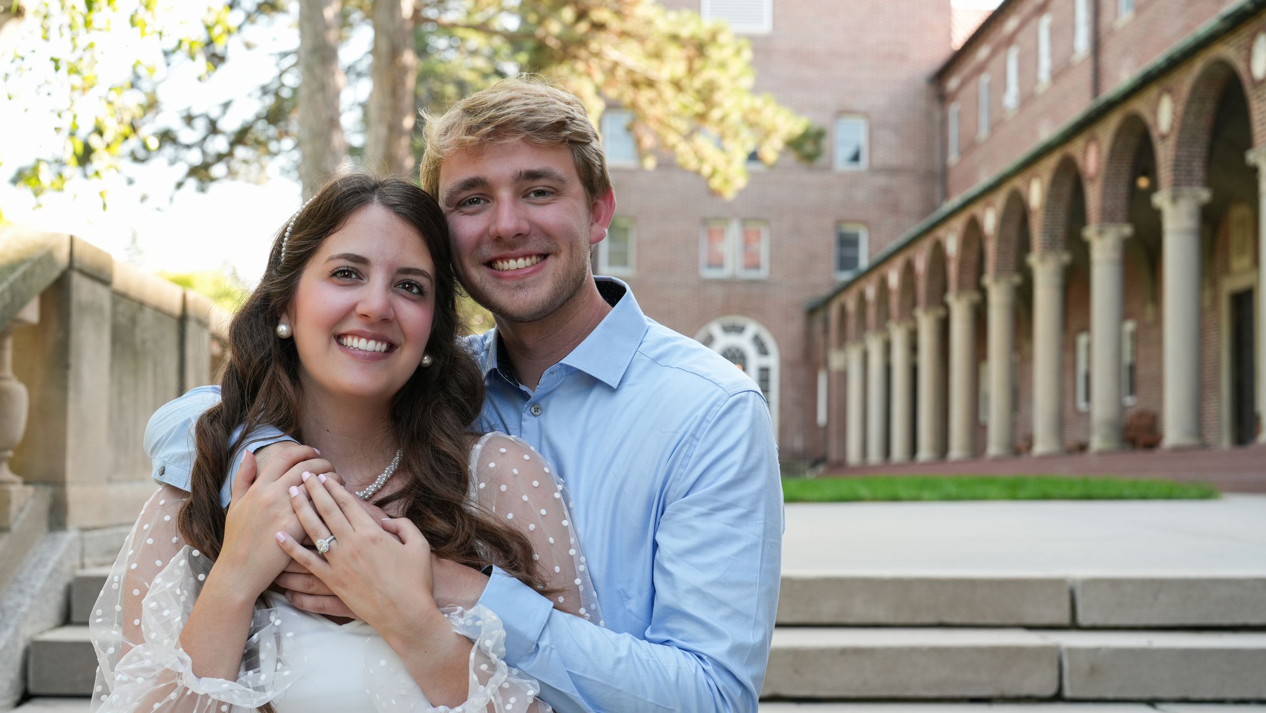 Smiling engaged couple posing together in front of a brick university building at sunset.