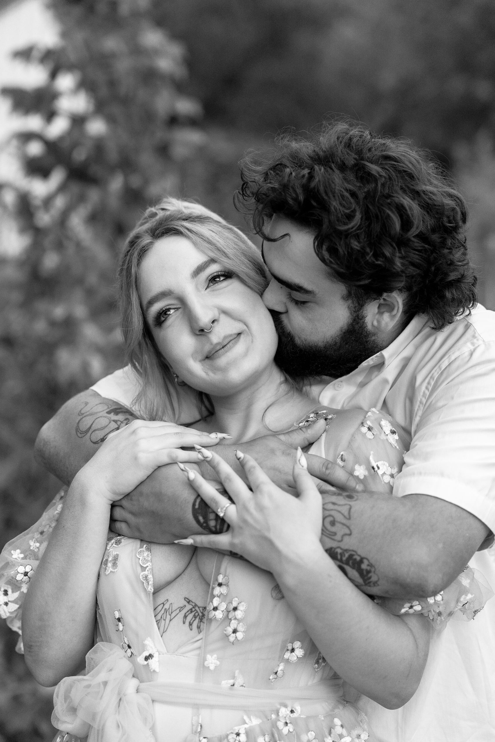 Black and white photo of a bearded man kissing a tattooed blonde woman outdoors in a romantic embrace.
